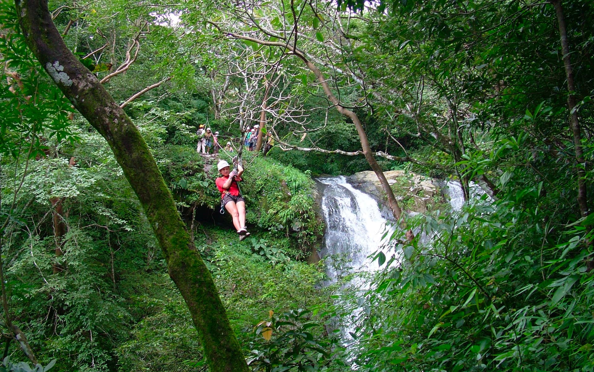 Person zip-lining near lush waterfall.