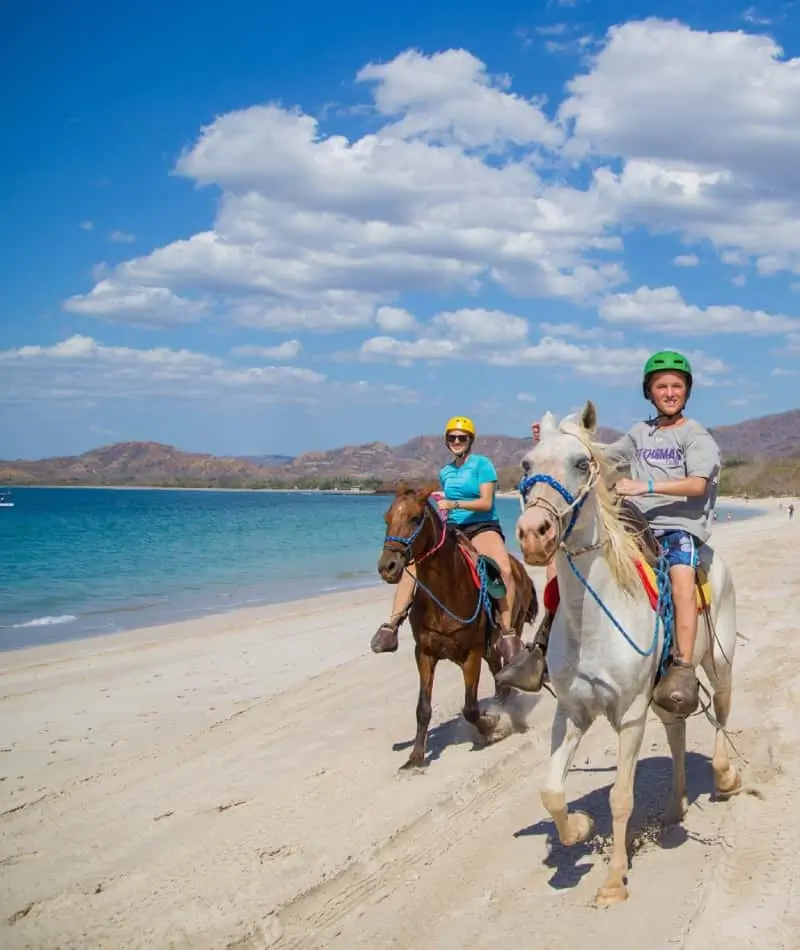 Two riders on horses by beach.